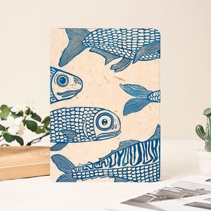 May include: A rectangular notebook with a beige background and a blue fish print. The fish are stylized with scales and fins, and are arranged in a repeating pattern. The notebook is a unique and artistic item.