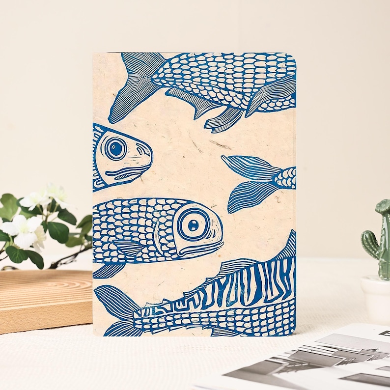 May include: A rectangular notebook with a beige background and a blue fish print. The fish are stylized with scales and fins, and are arranged in a repeating pattern. The notebook is a unique and artistic item.