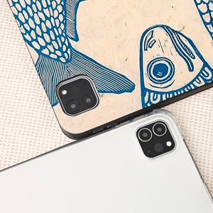 May include: Two tablet cases. One case features a blue fish design on a beige background. The other case is white. Both cases have a camera cutout with two lenses.