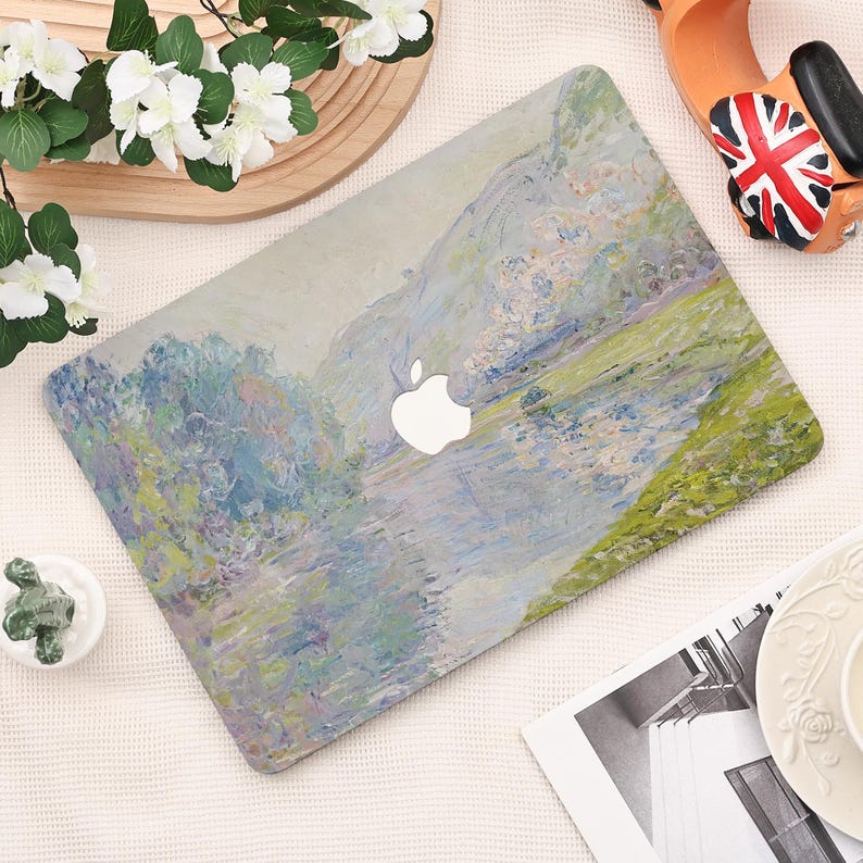 Pode incluir: Uma capa para laptop com um design de paisagem impressionista em tons de verde, azul e cinza. O logotipo da Apple &eacute; vis&iacute;vel. A capa est&aacute; sobre uma superf&iacute;cie branca com elementos decorativos.