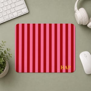 Puede incluir: Una alfombrilla de ratón rectangular con un patrón de rayas verticales que alterna tonos de rosa brillante y rojo oscuro. La esquina inferior derecha de la alfombrilla muestra el texto amarillo en mayúsculas HAP en una fuente serif simple.