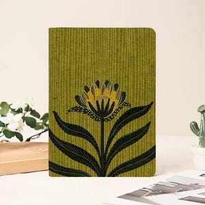May include: A green notebook with a floral design. The cover features a large black and yellow flower with long, dark green leaves against a textured, vertical-striped background. The notebook is rectangular and appears to be closed.
