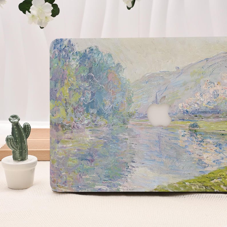 Pode incluir: Uma capa para laptop com uma pintura de paisagem apresentando um rio com &aacute;rvores e montanhas em tons de azul, verde e branco. O logotipo da Apple &eacute; vis&iacute;vel. Uma pequena estatueta de cacto em um vaso branco est&aacute; &agrave; esquerda.
