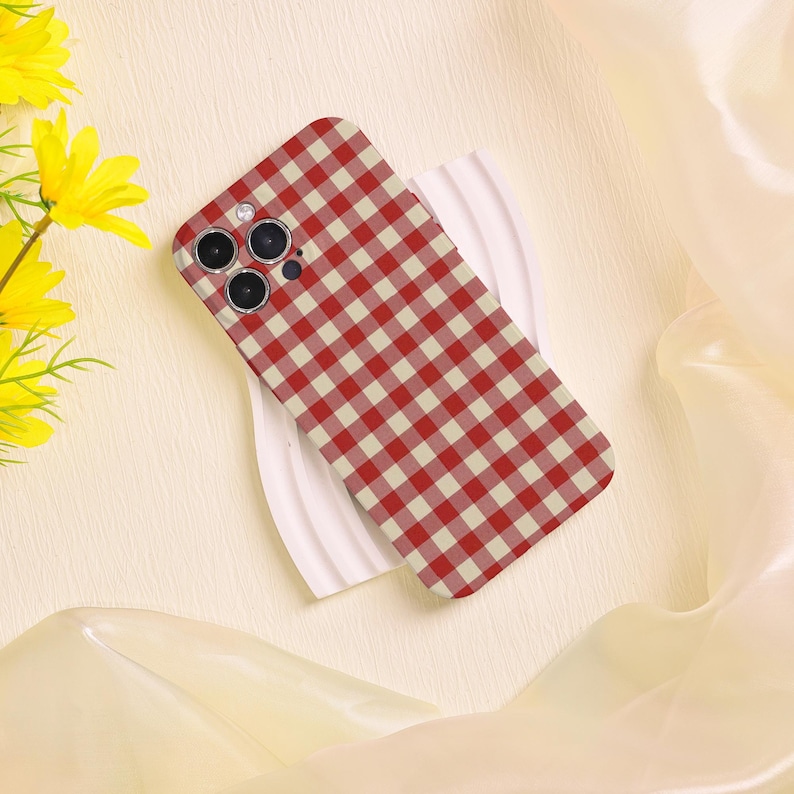 May include: A red and cream gingham patterned phone case. The case is resting on a white surface, with yellow flowers in the background. The phone case has three camera lenses.