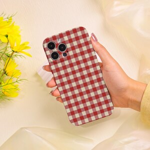 May include: A red and cream gingham pattern phone case. The case has a rectangular shape with rounded corners and a camera cutout. The case is held in a hand, with yellow flowers in the background.