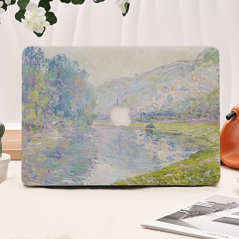 Pode incluir: Uma capa para laptop com uma pintura de paisagem retratando um rio, &aacute;rvores e uma montanha. A capa tem uma paleta de cores claras com tons de azul, verde e branco. O logotipo da Apple &eacute; vis&iacute;vel.