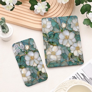 Może przedstawiać: Etui na telefon i etui na tablet z kwiatowym wzorem. Etui mają białe kwiaty ze złotymi środkami i łodygami na niebiesko-zielonym tle. Projekt przypomina witraż.
