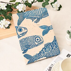 May include: A beige notebook with a blue fish print. The design features several fish illustrations in various poses, with detailed scales and fins. The word "FISH" is printed on the bottom right corner of the notebook.