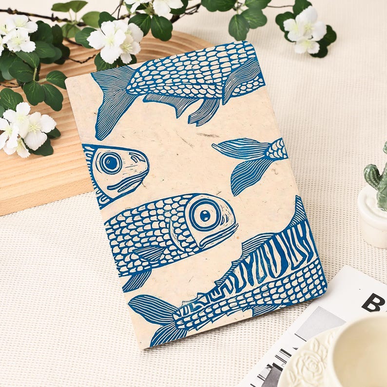 May include: A beige notebook with a blue fish print. The design features several fish illustrations in various poses, with detailed scales and fins. The word "FISH" is printed on the bottom right corner of the notebook.