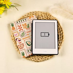 以下が含まれることがあります： 黒いフレームの白い電子書籍リーダーで、画面にはバッテリー残量低下のアイコンが表示されています。その隣には、パイナップル、バナナ、アボカド、花などのカラフルなイラストがクリーム色の背景に繰り返される模様の保護カバーが置かれています。