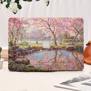 May include: A laptop case featuring a scenic painting of a pond surrounded by cherry blossom trees. The artwork depicts a serene landscape with a couple, swans, and koi fish. The case has a cutout for the Apple logo.