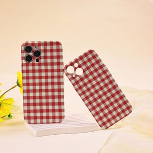 May include: Two red and white checkered phone cases. One case is upright, and the other is leaning against it. The cases are on a white platform with a yellow flower in the background. The cases have cutouts for the phone's camera.