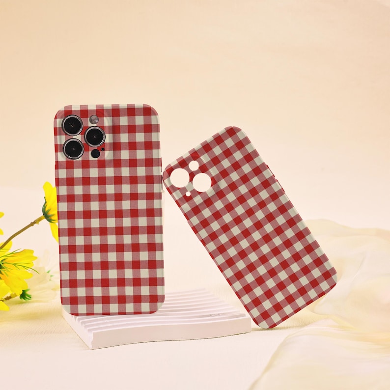May include: Two red and white checkered phone cases. One case is upright, and the other is leaning against it. The cases are on a white platform with a yellow flower in the background. The cases have cutouts for the phone's camera.