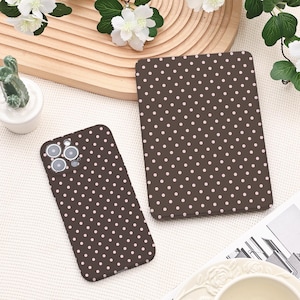 Op de afbeelding: Een bruine telefoonhoes en tablethoes met een herhalend patroon van kleine, lichtroze stippen. De telefoonhoes heeft een camera-uitsparing. De hoesjes worden gepresenteerd op een wit oppervlak met decoratieve elementen.