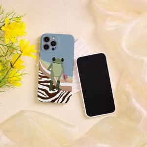以下が含まれることがあります： カウボーイブーツと帽子をかぶったカエルが描かれた青と白の携帯電話ケース。ケースにはゼブラ柄のデザインが施されています。