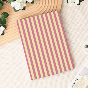 Op de afbeelding: Een rechthoekig notitieboek met een roze en gele streepjespatroon. De strepen lopen verticaal. Het notitieboek ligt op een wit oppervlak, met een houten dienblad en witte bloemen op de achtergrond.