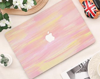 May include: A pink and yellow painted laptop cover with the Apple logo. The cover features a watercolour-style design with pink and yellow brushstrokes. The laptop cover is on a white surface with decorative items.