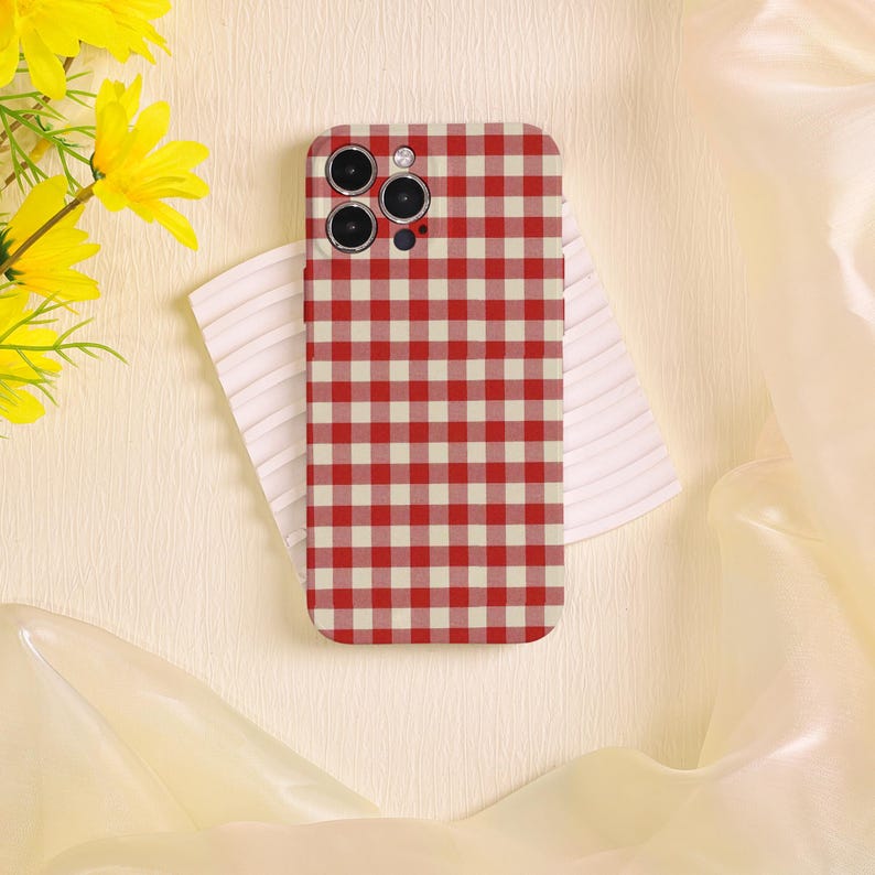 May include: A red and white checkered phone case with a triple-lens camera cutout. The case is resting on a white, textured surface, with yellow flowers in the upper left corner. The background is a soft, cream color.