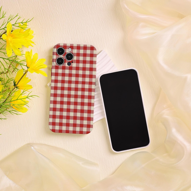 May include: A red and white checkered phone case and a black smartphone with a white border are displayed on a cream-colored surface. Yellow flowers are in the background. The phone case has a camera cutout.