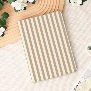 May include: A notebook with a beige and white striped pattern. The notebook is rectangular and lies on a white surface, with flowers and a wooden tray in the background. The cover design features vertical stripes.