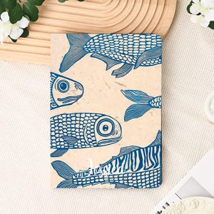May include: A rectangular notebook with a beige background and a blue fish print design. The fish are stylized with detailed scales and fins. The notebook is lying on a white surface.
