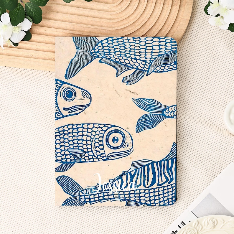 May include: A rectangular notebook with a beige background and a blue fish print design. The fish are stylized with detailed scales and fins. The notebook is lying on a white surface.