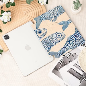 May include: A white iPad Pro with a clear case next to a beige case featuring a blue fish pattern. The fish design is a repeating pattern of stylized fish. The iPad is on a light-colored surface with decorative elements.