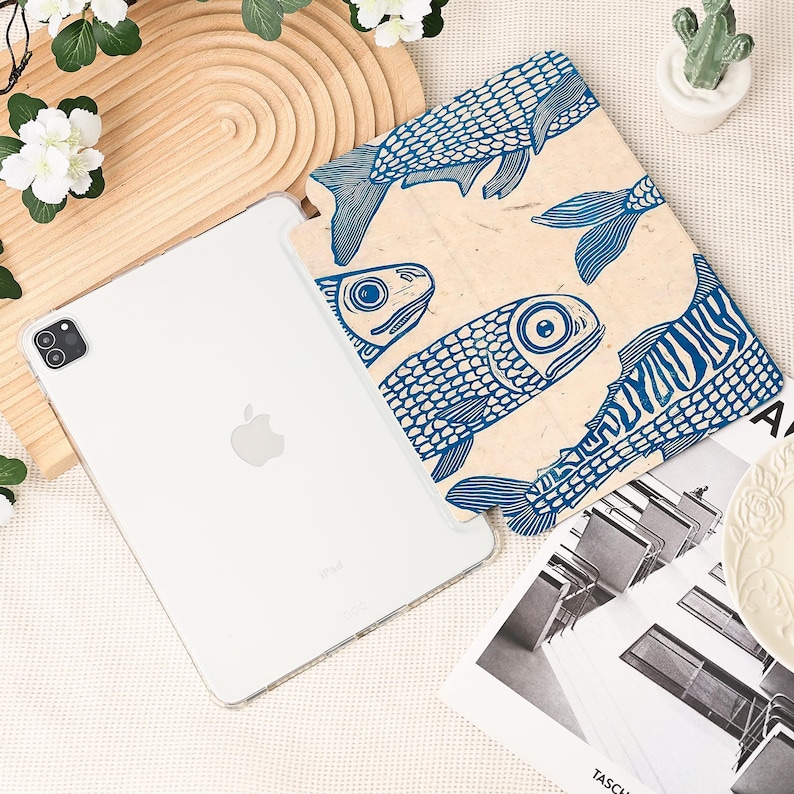 May include: A white iPad Pro with a clear case next to a beige case featuring a blue fish pattern. The fish design is a repeating pattern of stylized fish. The iPad is on a light-colored surface with decorative elements.