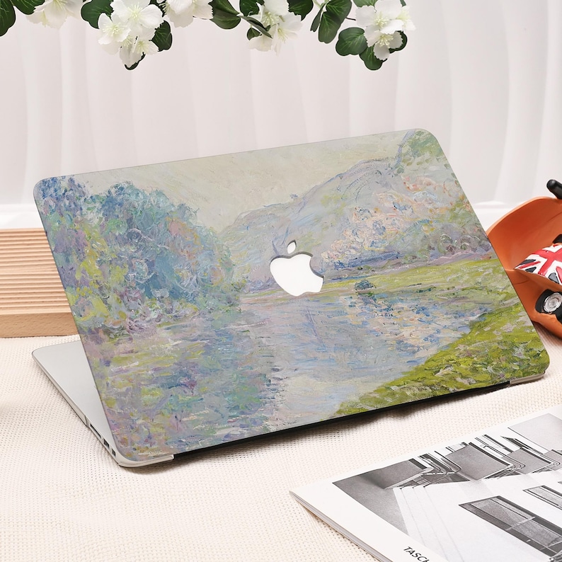 Pode incluir: Um laptop com um design de pintura de paisagem na capa. A obra de arte apresenta uma cena de rio com &aacute;rvores e montanhas, renderizada em suaves azuis, verdes e amarelos. O logotipo da Apple &eacute; vis&iacute;vel.