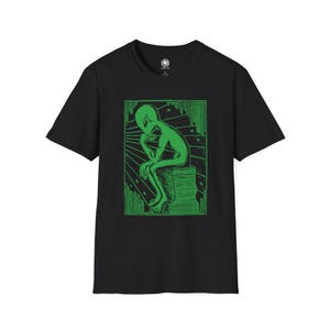以下が含まれることがあります： 緑色のグラフィックプリントが施された黒いTシャツ。プリントは、考え込んでいるポーズの宇宙人をモチーフにしている。
