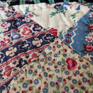 May include: A quilted textile featuring a patchwork design of various fabric patterns including blue and white floral prints, red and blue geometric stripes, a green and white gingham check, and a black and green plaid section with visible stitching.