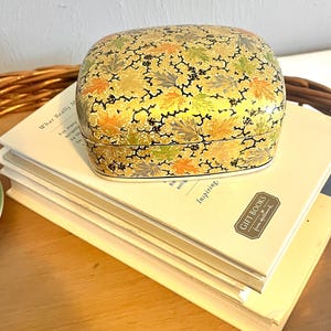 May include: A small, rectangular box with a colorful floral pattern. The box is made of paper mache and has a gold background with black, green, orange, and blue accents. The box is sitting on top of a stack of books.