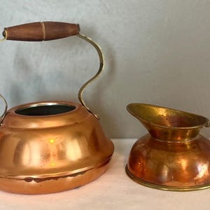 May include: A polished copper tea kettle with a dark brown wooden handle and a matching copper creamer pitcher. The kettle features a curved spout and a rounded body, while the creamer has a wide pouring lip and a curved brass-toned handle.