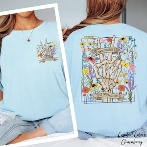 A woman wearing a light blue t-shirt with a graphic design on the back featuring a skeleton hand holding up a peace sign surrounded by flowers and the text "Good Energy is Contagious".