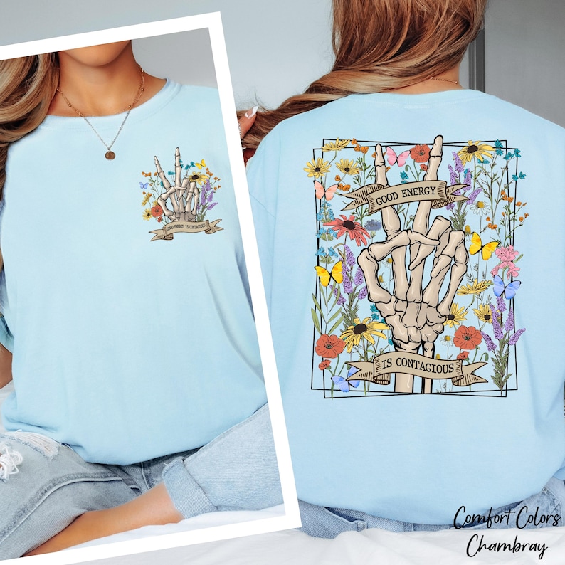 A woman wearing a light blue t-shirt with a graphic design on the back featuring a skeleton hand holding up a peace sign surrounded by flowers and the text "Good Energy is Contagious".