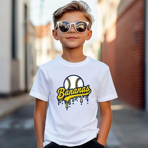 May include: White t-shirt with a baseball graphic and the word "Bananas" in yellow and blue. The baseball design has a blue outline and yellow stitching. The word "Bananas" has a dripping effect.