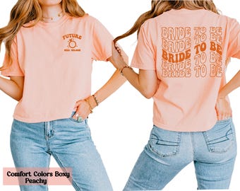 May include: Peach-colored t-shirt with "FUTURE MRS. WILSON" and a ring graphic on the front. The back of the shirt features the text "BRIDE TO BE" in a repeating, retro-style design. The shirt is paired with light blue jeans.