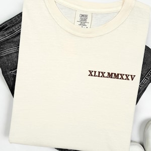 A white t-shirt with a brown embroidered text on it, along with a pair of black jeans and white shoes, all laid out on a white background.