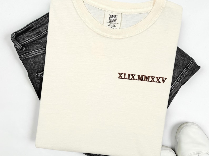 A white t-shirt with a brown embroidered text on it, along with a pair of black jeans and white shoes, all laid out on a white background.