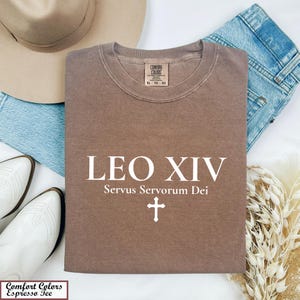 May include: A brown Comfort Colors Espresso Tee shirt with the text "LEO XIV Servus Servorum Dei" and a cross. The shirt is folded and surrounded by a hat, jeans, and white boots.