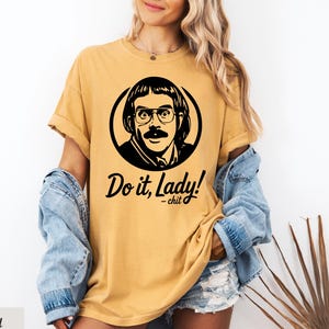 May include: Mustard yellow t-shirt featuring a black and white graphic of a man with glasses and a mustache inside a circle. The text "Do it, Lady! - chit" is below the graphic. The shirt has short sleeves and a relaxed fit.