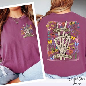 a woman wearing a purple shirt with a graphic design on the back. The design includes a skeleton hand making a peace sign, surrounded by flowers and butterflies. The text on the back of the shirt says "Good Energy is Contagious".