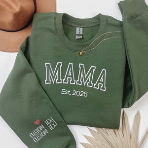 A green long-sleeved shirt with the word "MAMA" printed on it, along with the text "Est. 2025" below it. The shirt is placed on a white surface, with a brown hat and some leaves nearby.