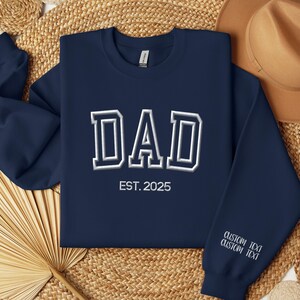 A navy blue sweatshirt with the text "DAD EST. 2025" printed on it, along with a straw hat and a fan, all laid out on a woven mat.