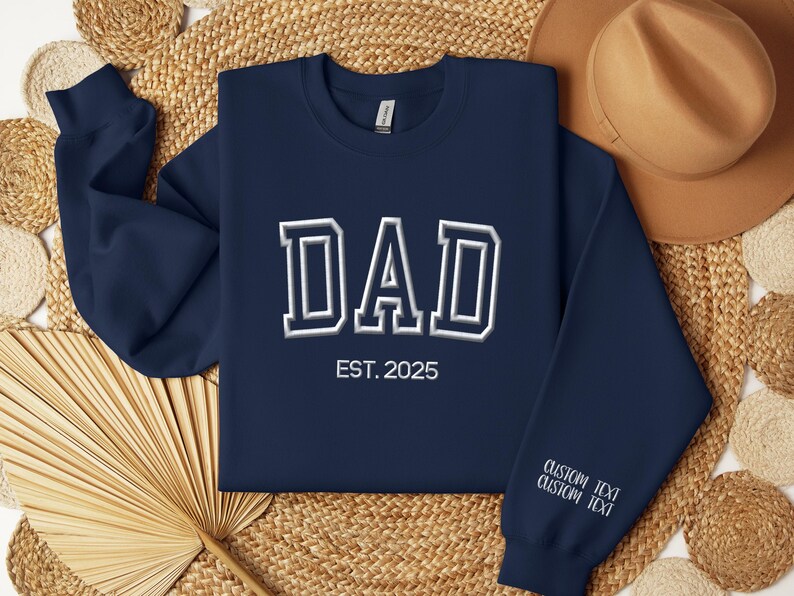 A navy blue sweatshirt with the text "DAD EST. 2025" printed on it, along with a straw hat and a fan, all laid out on a woven mat.