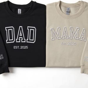 two long-sleeved shirts, one black and one beige, with the words "DAD" and "MAMA" printed on them. The shirts are laid out side by side, with the black shirt on the left and the beige shirt on the right.