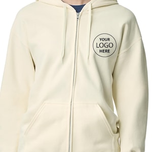 A person wearing a cream-colored zip-up hoodie with a black logo on the front.