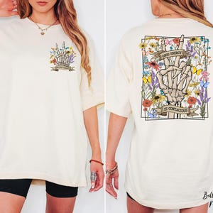 A woman wearing a white t-shirt with a graphic design on the back. The design features a skeleton hand holding up a peace sign, surrounded by flowers and a banner that reads "I am confident".