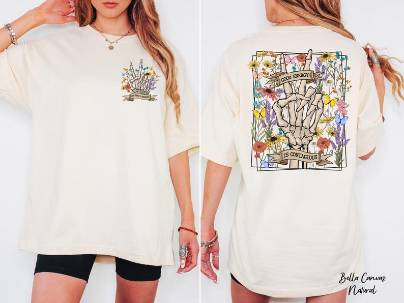 A woman wearing a white t-shirt with a graphic design on the back. The design features a skeleton hand holding up a peace sign, surrounded by flowers and a banner that reads "I am confident".
