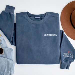 A blue sweatshirt with the text "XXIX MM XXV" printed on it, along with a brown hat and sunglasses, are arranged on a white background.
